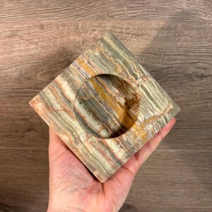 May include: Square ashtray made from layered onyx stone in shades of green, brown, and orange. The ashtray has a circular indentation in the centre. The ashtray is held in a hand.