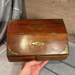 May include: A wooden box with brass accents and a hinged lid. The box has two brass plates with decorative designs on the front.