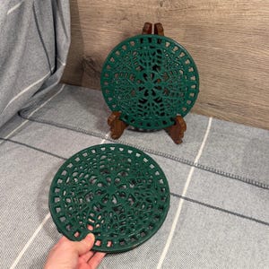 May include: Two round, dark green metal trivets featuring an intricate, openwork floral and geometric pattern. One trivet is displayed upright on a small wooden stand, while the other is held in the foreground, showing the detailed lattice design.