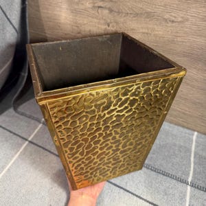 May include: A square wastebasket featuring a textured, hammered gold-tone metal exterior with a repeating organic pattern. The interior is dark and smooth, and the structure tapers slightly toward the base, showcasing a metallic finish throughout.