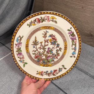 May include: A round ceramic plate with an off-white base featuring a central illustration of a flowering tree with pink, blue, and yellow blossoms. The rim is decorated with a brown geometric border and floral accents in pink, green, and blue.