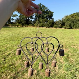 May include: A black metal wind chime featuring a double heart design. The wind chime has several small, aged bells hanging from the heart-shaped frame. The background is a green field and trees under a blue sky.