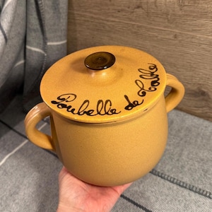 May include: A mustard-yellow ceramic pot with a lid and two handles. The lid has a dark brown knob and black cursive text that reads "Rubella de Lalle". The pot is round and has a textured surface.
