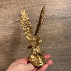 May include: A gold-coloured metal eagle figurine with its wings spread, perched on a rock-like base. The eagle is facing to the right, with its head turned back to look over its shoulder.