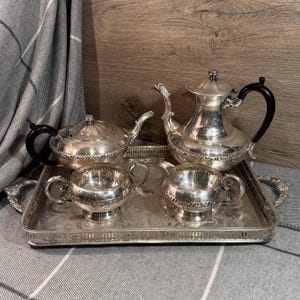May include: A silver tea set with black handles and a decorative tray. The set includes a teapot, a creamer, a sugar bowl, and a tea pot. The tray has ornate handles and a detailed border. The set is displayed on a grey and white patterned surface.