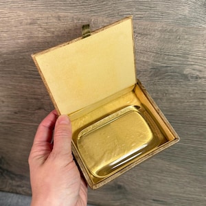 Op de afbeelding: Een vintage rechthoekige metalen tray in goudtinten in een bijpassende doos met een scharnierend deksel. De doos is gevoerd met een lichtgele stof. De tray heeft een licht gestructureerd oppervlak.