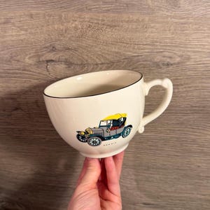 May include: A cream-colored ceramic mug with a dark rim and a handle, featuring a graphic illustration of a vintage car with a yellow roof and teal body. The word MORRIS is printed in small black letters beneath the car illustration on the mug.