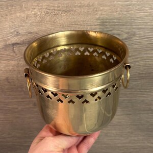 May include: Gold metal planter with heart-shaped cutouts. The planter has two handles on the sides.