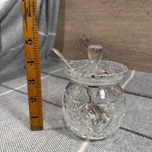 Op de afbeelding: Een heldere, geslepen glazen suikerpot met een deksel en een kleine lepel. De kom is ongeveer 7.6 cm hoog en heeft een ruitpatroon. De lepel is zilverkleurig. De kom staat op een grijs en wit gestreept oppervlak.