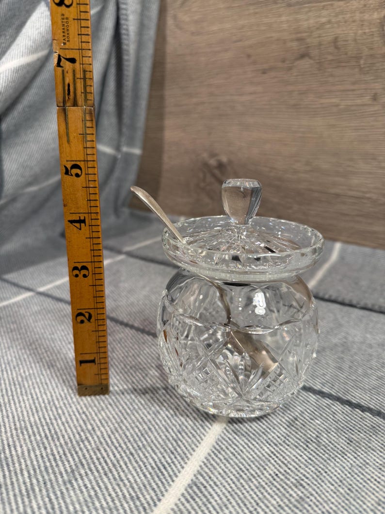 Op de afbeelding: Een heldere, geslepen glazen suikerpot met een deksel en een kleine lepel. De kom is ongeveer 7.6 cm hoog en heeft een ruitpatroon. De lepel is zilverkleurig. De kom staat op een grijs en wit gestreept oppervlak.