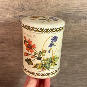 May include: Vintage metal canister with a cream-colored base and lid. The canister is decorated with floral illustrations in orange, purple, and blue. The text "Malva sylvestris" is written on the side. The rim of the lid and base feature a decorative gold and brown pattern.