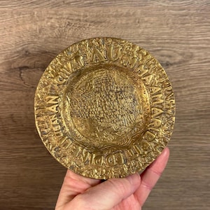 May include: A gold-coloured metal ashtray with a raised design of a city map and the text "EDINBURGH" and "AU MAA AMARIB" around the edge.