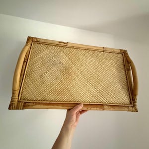 Puede incluir: Bandeja rectangular de bambú con centro tejido y asas curvas. La bandeja es de color marrón claro y está sujeta por la mano de una persona. El centro tejido tiene un patrón entrecruzado.