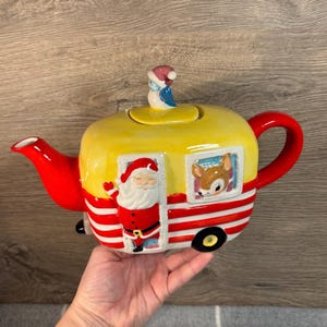 May include: A ceramic teapot shaped like a yellow and red camper van, featuring Santa Claus and a reindeer. The teapot has a red handle and spout, and a small penguin figurine on the lid.