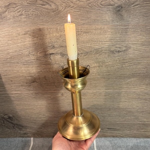 May include: A lit candle in a brass candlestick holder. The candle is off-white, and the holder has a round base, a long stem, and a small cup. The flame is bright, and the holder is held by a hand.