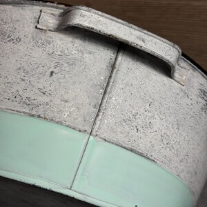 Op de afbeelding: Een ronde metalen bak met een verweerde witte afwerking aan de bovenzijde en een gladde, effen mintgroene band aan de onderzijde. De bak is voorzien van een gebogen metalen handvat dat aan de bovenrand is bevestigd met zichtbare naden.