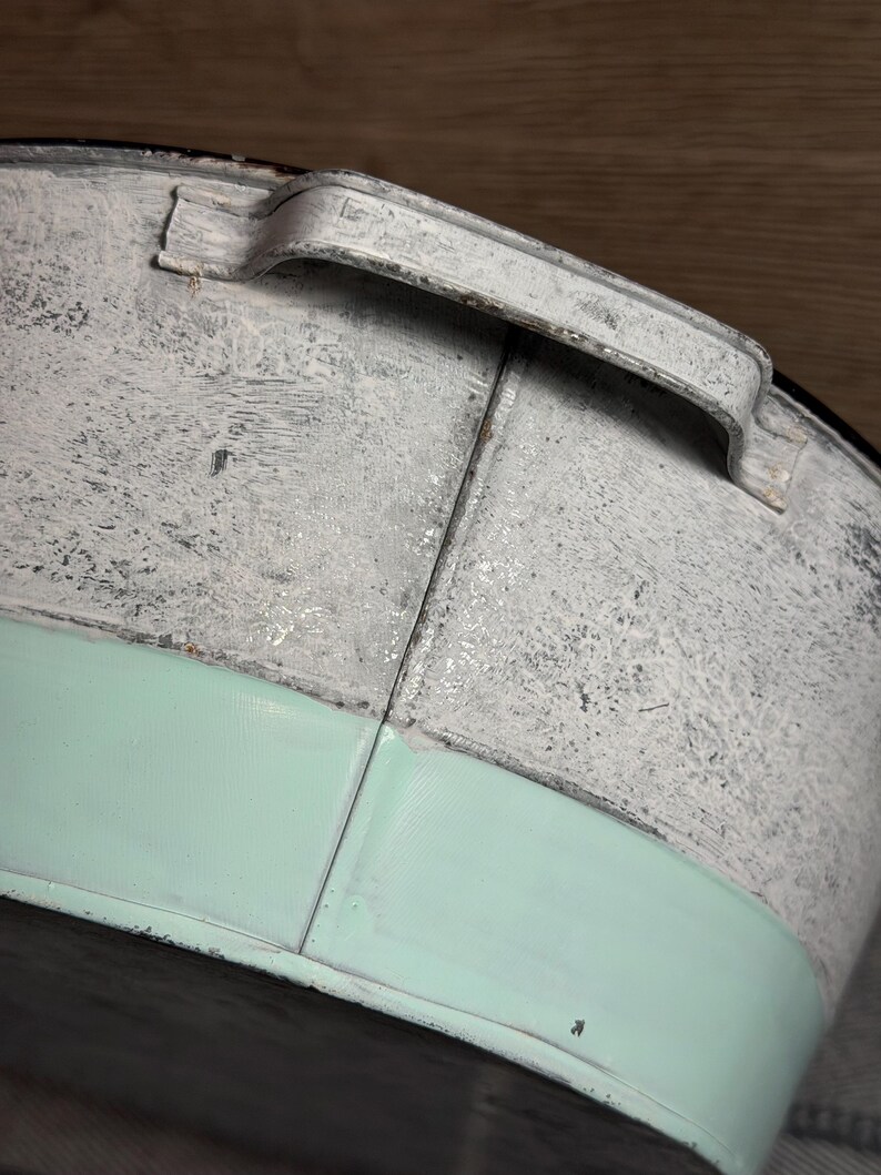 Op de afbeelding: Een ronde metalen bak met een verweerde witte afwerking aan de bovenzijde en een gladde, effen mintgroene band aan de onderzijde. De bak is voorzien van een gebogen metalen handvat dat aan de bovenrand is bevestigd met zichtbare naden.