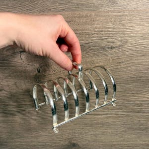 Puede incluir: Un tostador plateado vintage con seis ranuras curvas. El tostador tiene un asa en la parte superior y está hecho de metal. El tostador está diseñado para sostener rebanadas de tostadas en posición vertical. El fondo es una superficie de madera.