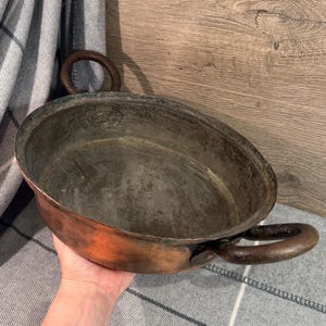 May include: An antique copper pan with two large, circular handles. The pan has a wide, shallow bowl with a dark interior and a reddish-brown exterior. The handles are a darker, aged metal. The pan is likely used for cooking.
