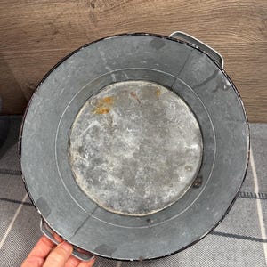 Op de afbeelding: Een ronde kom van gegalvaniseerd metaal met een donkere, verweerde grijze afwerking met zichtbare tekenen van oxidatie en roestvlekken op de binnenbodem. De kom is voorzien van twee stevige metalen handgrepen aan weerszijden van de rand.