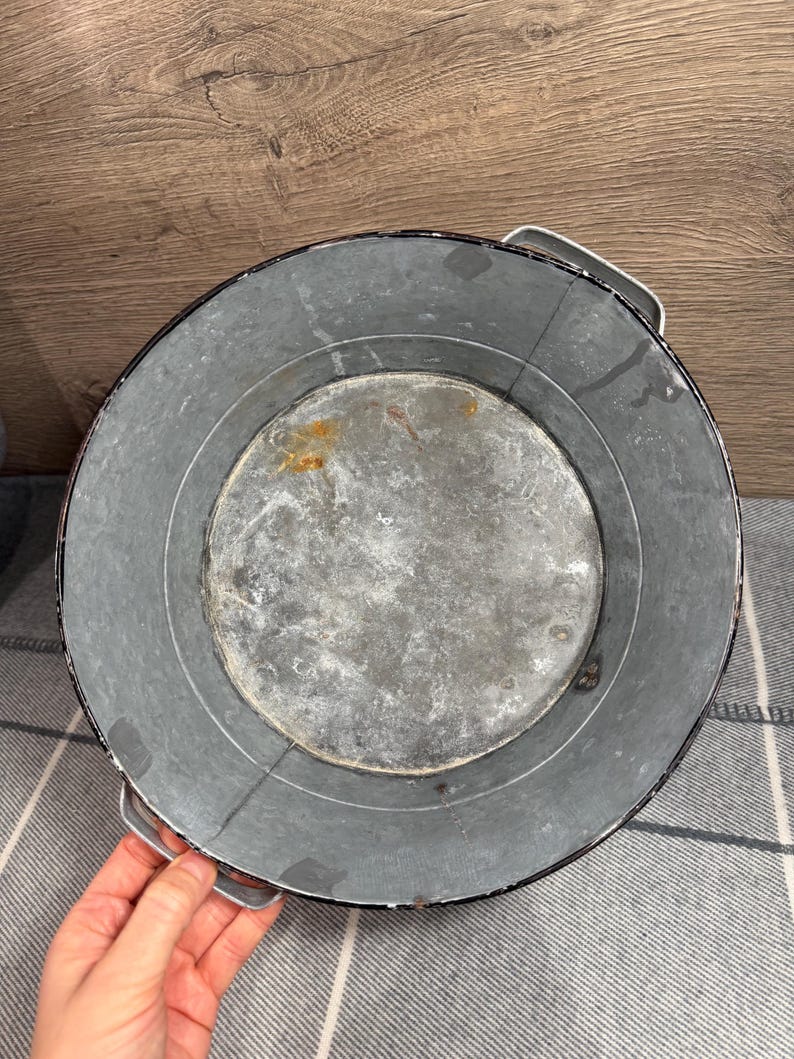 Op de afbeelding: Een ronde kom van gegalvaniseerd metaal met een donkere, verweerde grijze afwerking met zichtbare tekenen van oxidatie en roestvlekken op de binnenbodem. De kom is voorzien van twee stevige metalen handgrepen aan weerszijden van de rand.