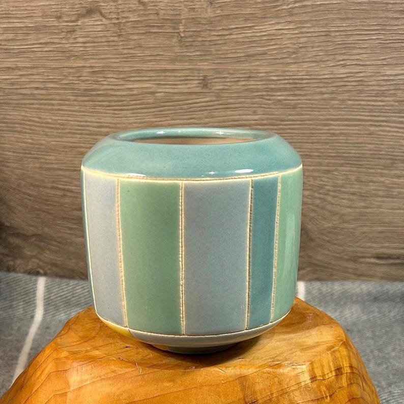 Op de afbeelding: Een kleine, cilindrische keramische pot met een lichtblauwe glazuur en verticale strepen in een lichter blauw en groen. De pot staat op een houten oppervlak.