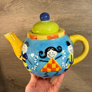 Op de afbeelding: Een grillige keramische theepot met een blauwe body, gele tuit en handvat en een groen deksel. De theepot is versierd met cartoonfiguren en een ruitpatroon. Het deksel heeft een blauwe knop.