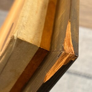 Puede incluir: Primer plano de un marco de madera con una esquina da&ntilde;ada. El marco es de color marr&oacute;n oscuro con una veta de madera marr&oacute;n m&aacute;s clara visible. La esquina da&ntilde;ada est&aacute; astillada y muestra la madera m&aacute;s clara debajo.