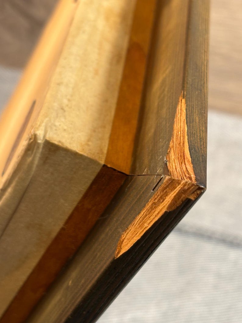 Puede incluir: Primer plano de un marco de madera con una esquina da&ntilde;ada. El marco es de color marr&oacute;n oscuro con una veta de madera marr&oacute;n m&aacute;s clara visible. La esquina da&ntilde;ada est&aacute; astillada y muestra la madera m&aacute;s clara debajo.