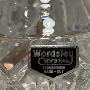 Op de afbeelding: Close-up van een helder kristallen glas met een diamantgeslepen patroon. Een zwart en zilveren label leest "Wordsley Crystal, Stourbridge, Hand-Cut."