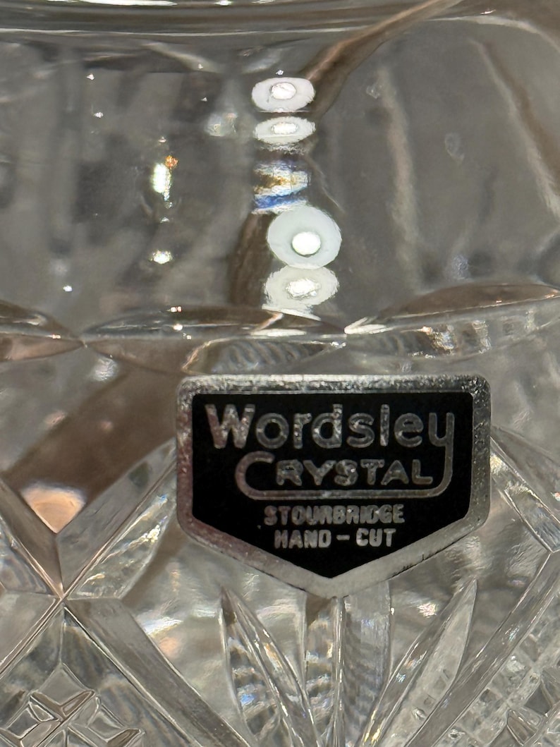 Op de afbeelding: Close-up van een helder kristallen glas met een diamantgeslepen patroon. Een zwart en zilveren label leest "Wordsley Crystal, Stourbridge, Hand-Cut."