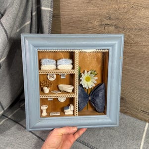 May include: A light blue wooden shadow box frame containing a miniature bathroom scene. The interior features shelves with tiny white towels, a bathtub, a toilet, a sink, a potted plant, a white daisy flower, and a large blue paper bow.