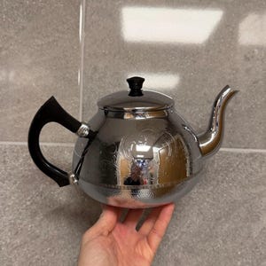 May include: A shiny silver teapot with a black handle and lid. The teapot features a curved spout and an etched floral design. The teapot is held in a person's hand.