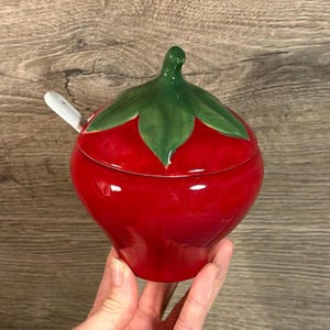 Puede incluir: Un azucarero de cerámica rojo con forma de fresa, con un diseño de hoja verde y una tapa blanca con una pequeña cuchara.