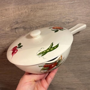 Puede incluir: Plato de cerámica blanca para hornear con tapa y asa. El plato está decorado con una vaina de guisantes verdes, rábanos rojos y un tomate rojo. La tapa tiene un pomo en el centro.