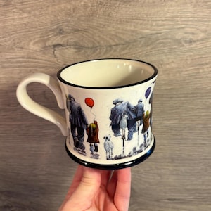 以下が含まれることがあります： 黒い縁と持ち手が付いた白いセラミックマグ。犬と歩く人々と赤い風船を持った子供のイラストが描かれています。マグカップは青いベースと白い内側を持っています。