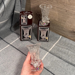 Puede incluir: Floreros de cristal transparente con intrincados diseños florales y de estrellas. Los floreros tienen una parte superior acampanada y una base decorativa. Dos floreros se muestran en sus cajas originales, y uno se sostiene en una mano. Las cajas son marrones oscuras.