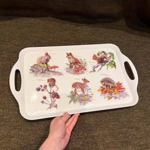 Puede incluir: Una bandeja de servicio rectangular blanca con asas integradas que presenta seis ilustraciones detalladas de animales del bosque, incluyendo un conejo, un zorro, una ardilla, un ratón, un cervatillo y un erizo, rodeados de elementos naturales.