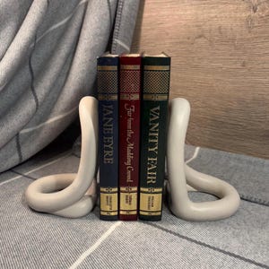 Peut inclure: Deux serre-livres en céramique blanche en forme de boucle continue, tenant trois livres reliés. Les livres sont intitulés "Jane Eyre", "Loin de la foule déchaînée" et "La foire aux vanités".