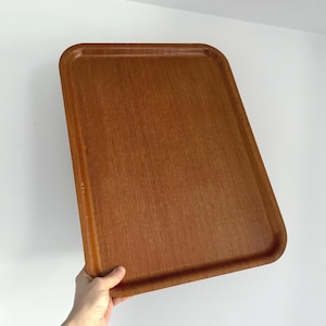 May include: A rectangular wooden serving tray with rounded corners. The tray is a warm brown colour with a visible wood grain texture. The tray is being held by a hand, showing its size and shape. The tray is designed for serving or carrying items.