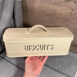 Może przedstawiać: Kremowa, prostokątna puszka na herbatniki z uchwytem na górze. Słowo "BISCUITS" jest wycięte z przodu. Puszka jest trzymana przez dłoń, z szaro-białym kocem w tle.
