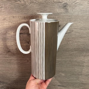 Peut inclure: Une cafetière en céramique blanche avec un couvercle, une anse et un bec verseur. Le corps est divisé en deux sections verticales, l'une avec des lignes verticales blanches et marron, l'autre avec des lignes marron. La cafetière est tenue par une main.