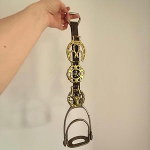 Op de afbeelding: Een bruin leren paardentuig met drie decoratieve messing medaillons en een zilveren stijgbeugel. De medaillons hebben hoefijzer-, cirkel- en hertenmotieven. Het tuig wordt omhoog gehouden door een zilveren metalen lus.