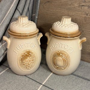 May include: Two ceramic jars with lids, both with the word "Rumtopf" written on them. The jars are white with brown accents and feature a design of a fruit and leaves.