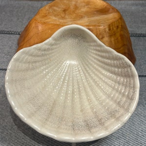 Op de afbeelding: Een wit keramisch schaaltje in de vorm van een schelp met een gekrakelde glazuur. Het schaaltje staat op een houten voet.
