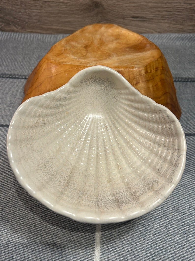 Op de afbeelding: Een wit keramisch schaaltje in de vorm van een schelp met een gekrakelde glazuur. Het schaaltje staat op een houten voet.