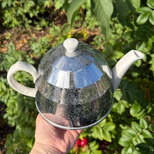 Könnte beinhalten: Eine reflektierende, silberfarbene Teekanne mit weißem Griff, Ausguss und Deckel. Die Teekanne hat eine runde Form und eine strukturierte Oberfläche. Der Hintergrund ist ein grüner Garten. Durchmesser ca. 20 cm.