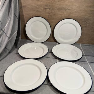 Pode incluir: Um conjunto de seis pratos redondos de esmalte branco dispostos em duas colunas de tr&ecirc;s. Cada prato apresenta uma superf&iacute;cie branca lisa e brilhante com uma borda azul escura distinta ao longo do contorno. Os pratos s&atilde;o uniformes no design.
