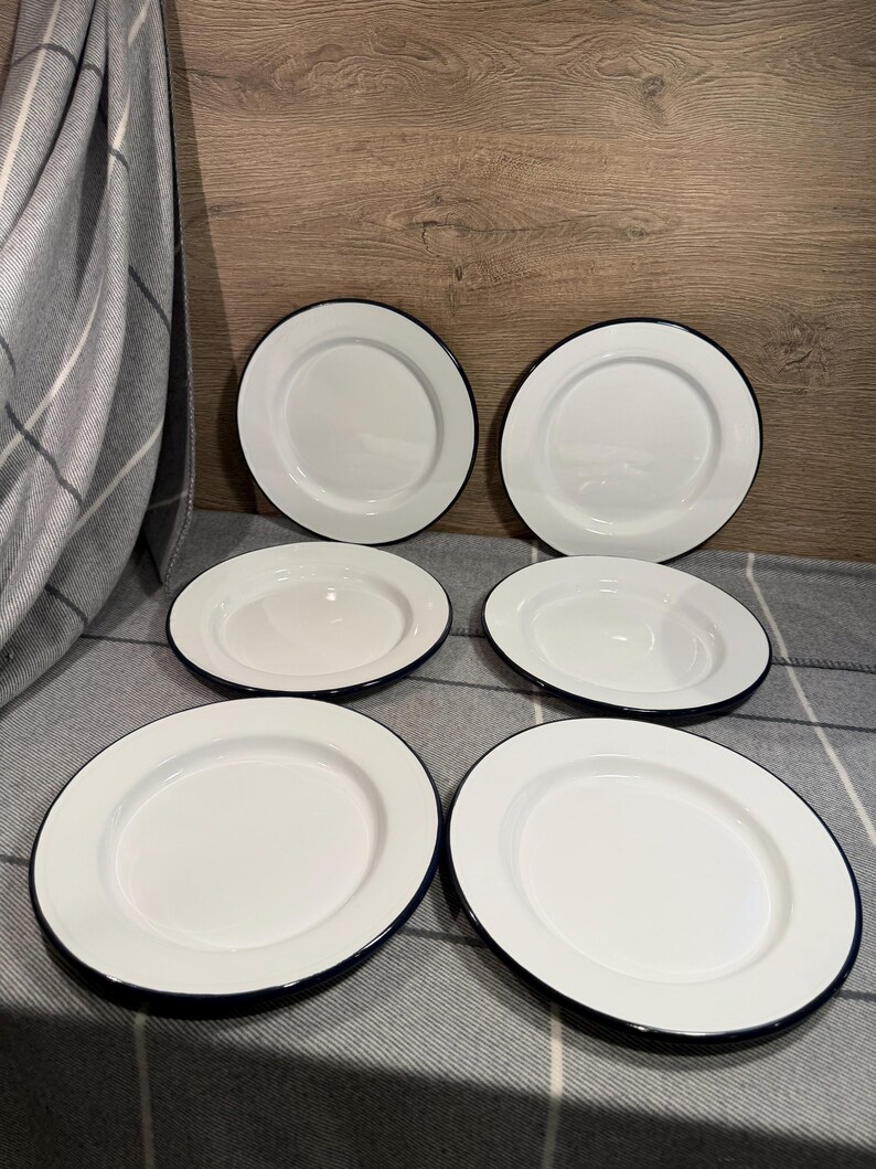 Pode incluir: Um conjunto de seis pratos redondos de esmalte branco dispostos em duas colunas de tr&ecirc;s. Cada prato apresenta uma superf&iacute;cie branca lisa e brilhante com uma borda azul escura distinta ao longo do contorno. Os pratos s&atilde;o uniformes no design.