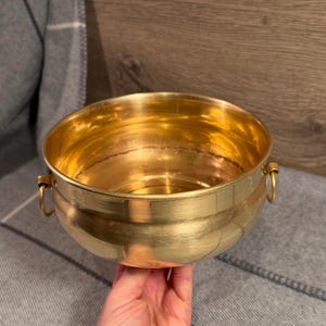 May include: A shiny, gold-colored metal bowl with two small ring handles. The bowl has a rounded shape and a smooth surface. The bowl is held by a person.